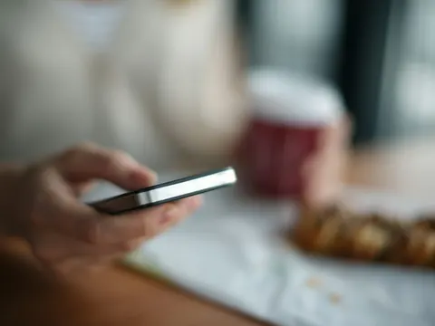 Hand mit Smartphone am Tisch, daneben Kaffeebecher und Gebäck